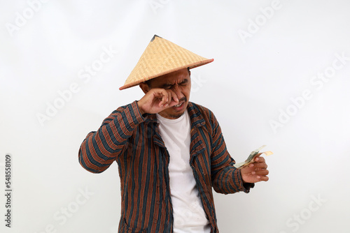 Disappointed asian farmer standing while holding little money and crying. Isolated on white background