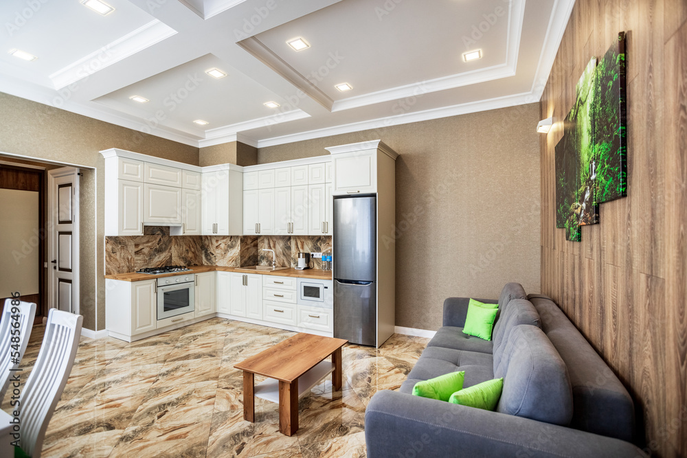 studio kitchen in apartments in a modern style. white kitchen with wooden countertop. White dining table with white chairs. Brown sofa.