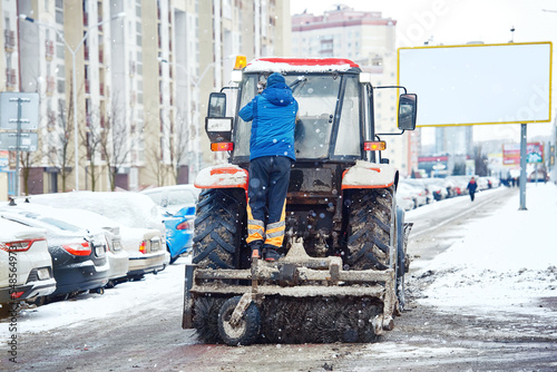 Tractor with brush and scraper removes snow at city street, clears sidewalk during heavy snowfall at winter day. Tractor clearing snowy streets with snowplough and rotary brush. Vehicle sweeps snow