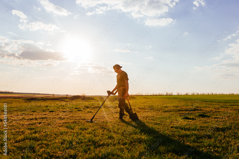 Fototapeta premium Treasure hunter stands against the blue sky. Treasure hunter holding a modern metal detector. Guy with a shovel looking for treasure