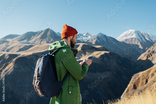 a man with a backpack in a green raincoat stands and looks at the phone in the mountains