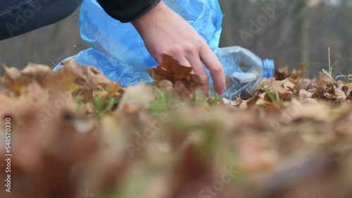 A man picking up a plastic bottle and putting it in a blue garbage bag in the park. The concept of plastic pollution, forest cleaning. 