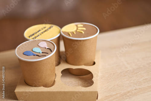 three homemade mini chocolate cake to go in coffee cup with cream drawing on a table, new confectionery trend concept. Paper cups with cakey are in a box. fashionable dessert. Horizontal, copy space