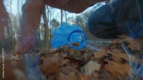 A man picking up a plastic bottle and putting it in a blue garbage bag in the park, pov view from inside of the bottle. The concept of plastic pollution, forest cleaning. 
