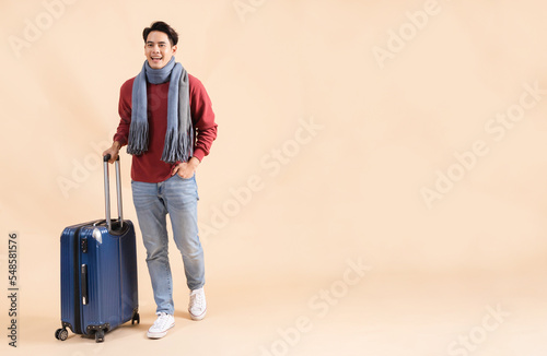 Happy Asian male tourist with luggage in red sweater and scarf enjoying winter vacation isolated on beige color background. Excited fashionable 20s guy going on a trip - Full body length studio shot.