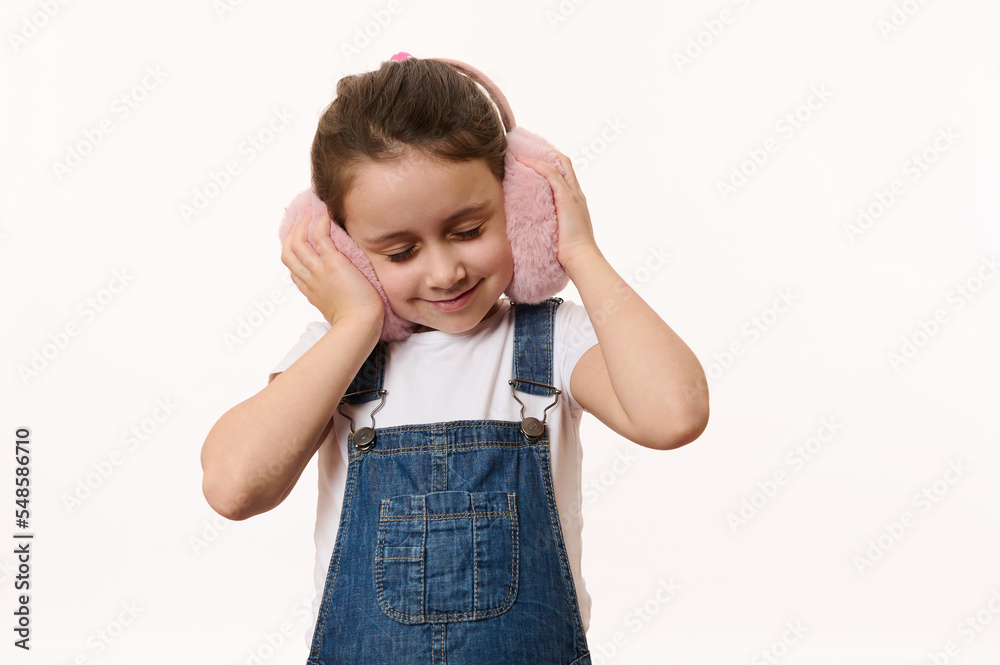 Fashionable little girl in blue denim overalls, holding her hands on furry plush earmuffs, enjoying her new stylish winter accessory. Protect your ears from the cold, isolated over white background Fashionable little girl in blue denim overalls, holding her hands on furry plush earmuffs, enjoying her new stylish winter accessory. Protect your ears from the cold, isolated over white background