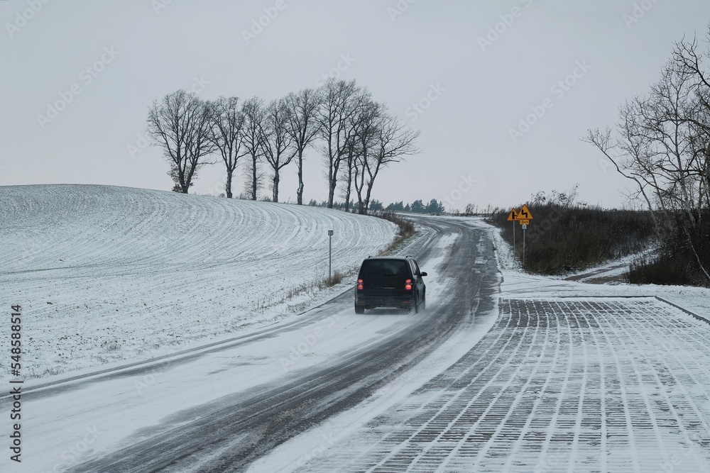 Car on asphalt road among fields in heavy road conditions with snow on slippery surface