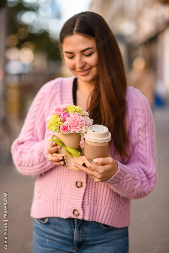 Fototapeta premium pretty woman gently holds flower arrangement in small pot and paper cup of coffee in her hands.