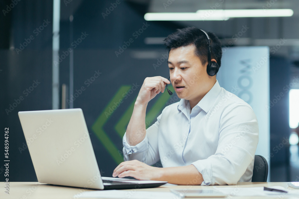 Fototapeta premium Portrait of young Asian male consultant in support service. Sitting in the office at the table, wearing headphones and a microphone, talking with clients online from a laptop.