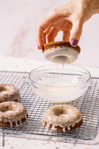 A female dipping a doughnut into white icing in a bowl, and decorated doughnuts on the cooking rack