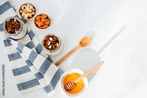 Yoghurts in glass jars with granola, chocolate, honey and nuts on a white background with kitchen towel. The concept of healthy eating. Flat lay