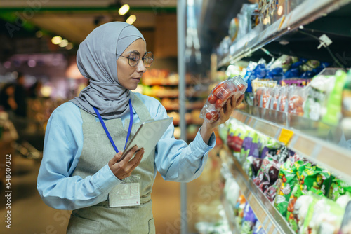 A Muslim woman in a hijab, a supermarket worker checks products on shelves and in refrigerators, a woman with a tablet and an apron uses a tablet to review products.