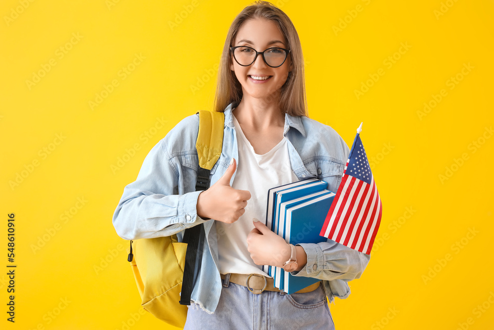 Beautiful woman with USA flag, books and backpack showing thumb-up on yellow background Beautiful woman with USA flag, books and backpack showing thumb-up on yellow background