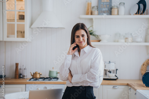 Stunning Brunette young hispanic woman in white shirt and black pants standing at kitchen looks at camera smiles touching face. Confident Caucasian female entrepreneur at home. Successful people.
