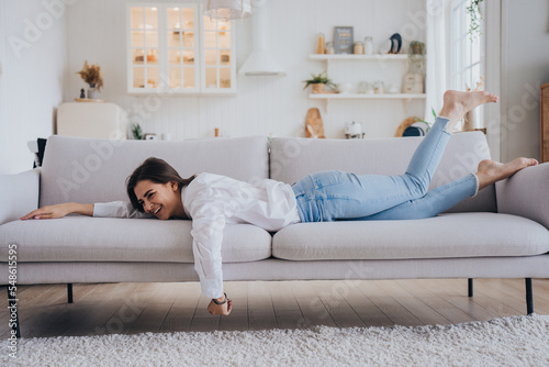 Fotografia Playful young brunette hispanic woman in white shirt and blue denim jeans laying on cozy sofa laughing having fun