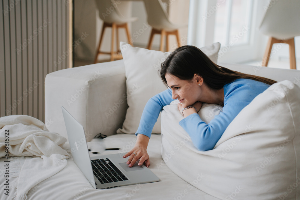 Young hispanic woman in blouse, jeans laying on sofa in front of laptop, leans on a large pillow, looks dreamily uses touch pad. Girl at video call full relaxing, education and remote work. Cozy home. Young hispanic woman in blouse, jeans laying on sofa in front of laptop, leans on a large pillow, looks dreamily uses touch pad. Girl at video call full relaxing, education and remote work. Cozy home.