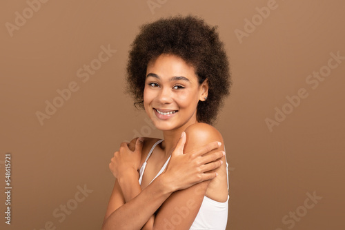 Cheerful millennial african american curly lady in white top with perfect skin and nude makeup hug her shoulders