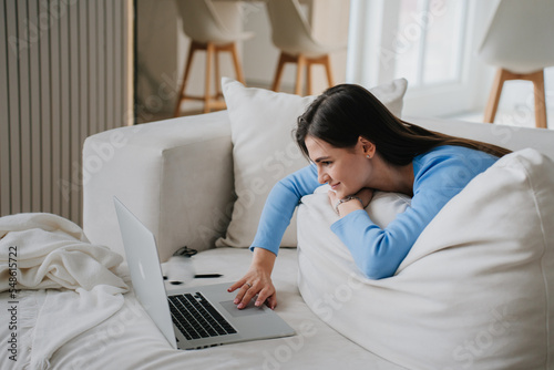 Young hispanic woman in blouse, jeans laying on sofa in front of laptop, leans on a large pillow, looks dreamily uses touch pad. Girl at video call full relaxing, education and remote work. Cozy home.