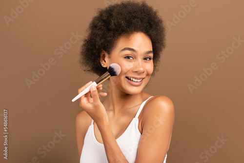 Cheerful millennial african american curly woman in white top applying blush or powder on face with brush