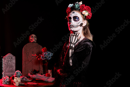 Spooky lady of death wearing black skull costume in studio, looking like holy santa muerte with flowers crown. Acting creepy and posing dressed as goddess of dead on dios de los muertos.