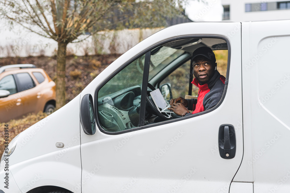 Hard-working focused Black delivery man checking the address and crossing it off of digital lis placed on his laptop. White delivery truck. Outdoor view. High quality photo Hard-working focused Black delivery man checking the address and crossing it off of digital lis placed on his laptop. White delivery truck. Outdoor view. High quality photo