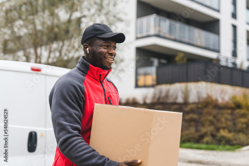 Technology in the package delivery business. Medium closeup portrait of a bearded handsome post office deliveryman delivering parcel packed in cardboard. Blurred modern building block in the