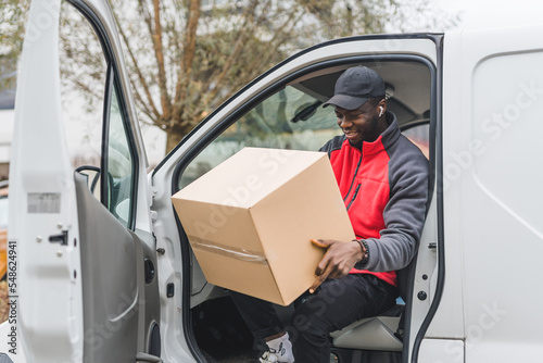 Black young adult delivery guy in work uniform sitting in door of white van looking at cardboard box parcel. Horizontal outdoor shot. High quality photo