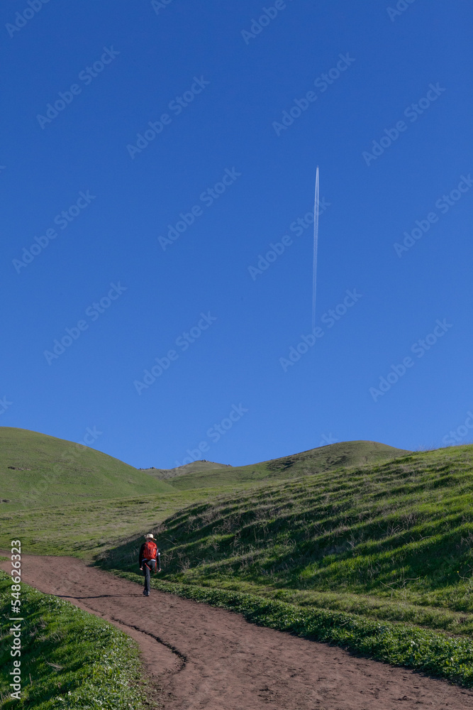 Obraz premium Female hiker walking up steep trail with con trail in the distance on a clear day at Mission Peak Park in Fremont, California