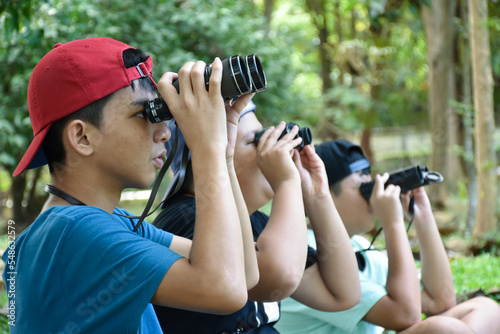 Asian boys are using binoculars to do the birds' watching in tropical forest during summer camp, idea for learning creatures and wildlife animals and insects outside the classroom.