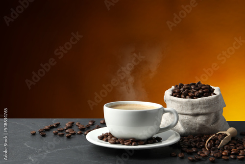 Cup of hot aromatic coffee and roasted beans on black table against brown background. Space for text
