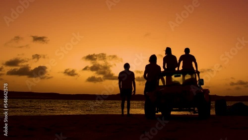 Silhouette showing group of people with quad watching sunset and jumping kiteboarder in ocean - static wide shot