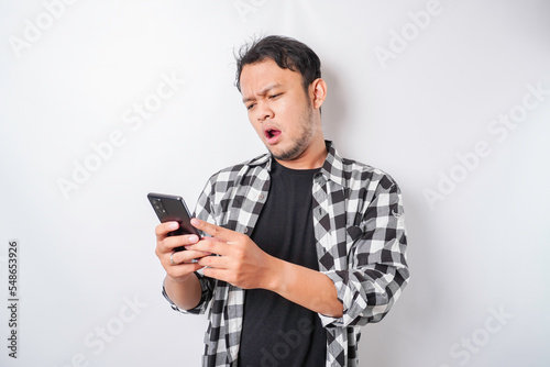 A dissatisfied young Asian man looks disgruntled wearing tartan shirt irritated face expressions holding his phone