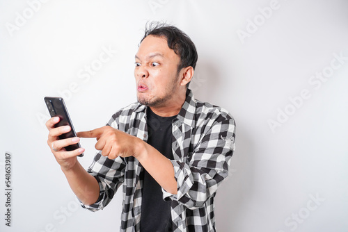 The angry and mad face of Asian man in tartan shirt while holding his phone on isolated white background. © Reezky