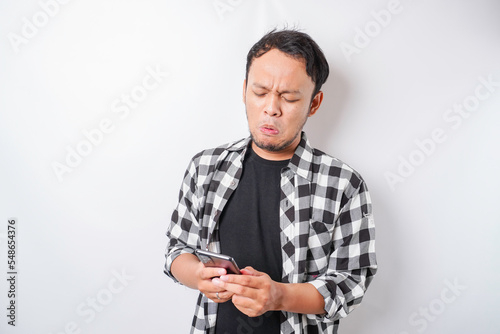 A dissatisfied young Asian man looks disgruntled wearing tartan shirt irritated face expressions holding his phone