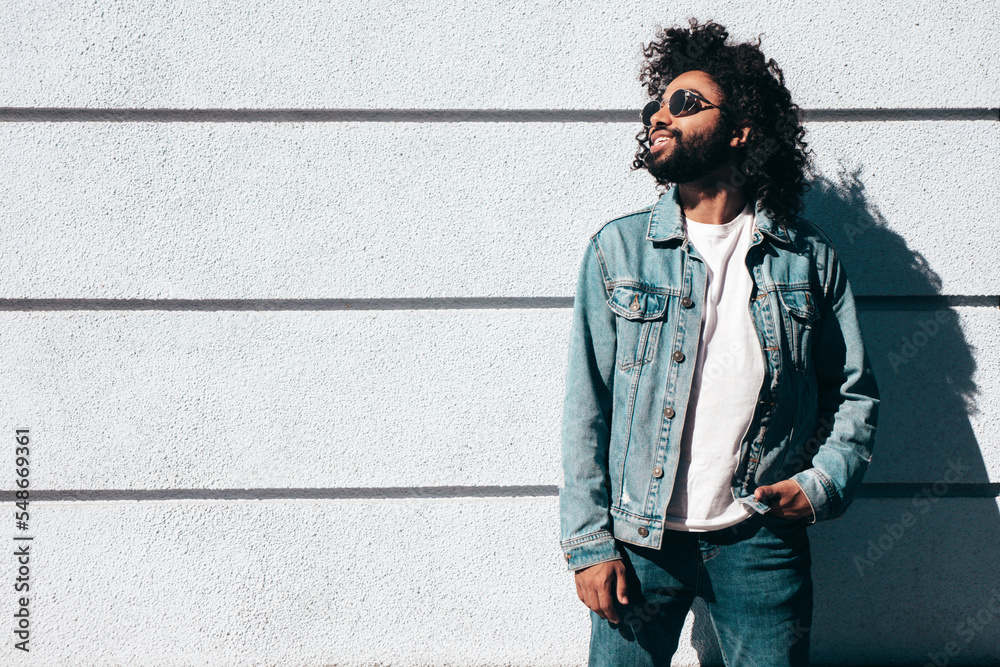Handsome smiling hipster  unshaven Arabian man dressed in summer  jeans jacket clothes. Fashion male with long curly hairstyle posing in  street at sunset. Cheerful and happy. In sunglasses Photos | Adobe