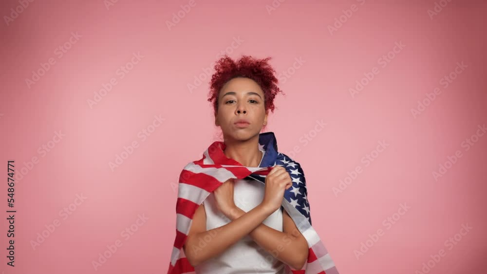 Serious African American patriot redhead curly woman drapes US flag over shoulders looks camera. Portrait female standing with USA flag on isolated pink background. 4th July America independence day Serious African American patriot redhead curly woman drapes US flag over shoulders looks camera. Portrait female standing with USA flag on isolated pink background. 4th July America independence day