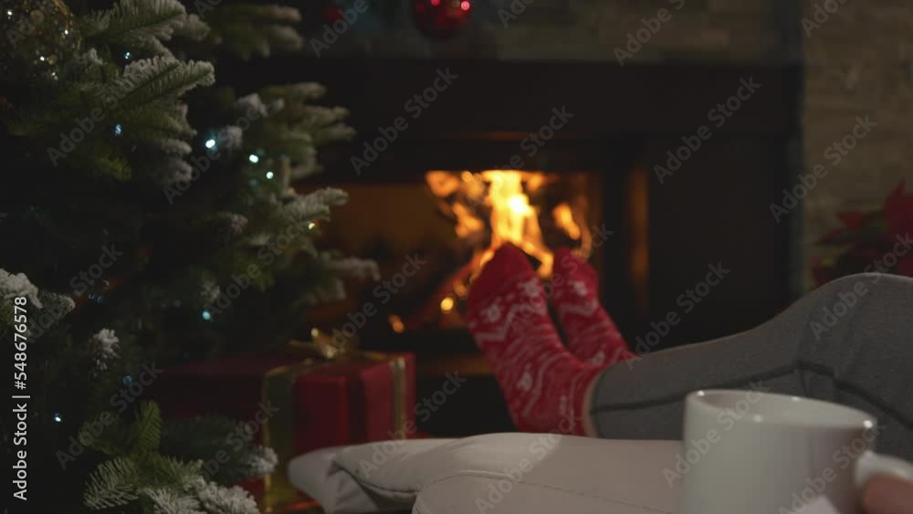 CLOSE UP: Woman has a relaxing evening drinking hot tea by the burning fireplace. Female person enjoying and resting in pleasant ambiance of her festively decorated home living room on Christmas Eve. CLOSE UP: Woman has a relaxing evening drinking hot tea by the burning fireplace. Female person enjoying and resting in pleasant ambiance of her festively decorated home living room on Christmas Eve.
