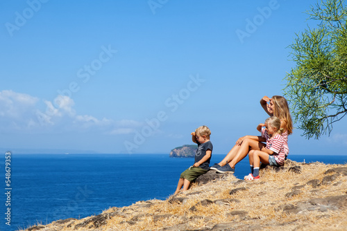 Happy mother with kids walking on summer sea beach holiday. Beautiful family relaxing on high ocean cliff. Looking at beautiful sky and sea. Healthy family lifestyle, summer travel on tropical island.