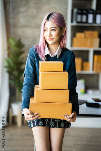 Young Asian SMEs woman holding parcels, standing among several boxes in modern office. © NINENII
