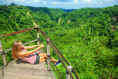 Family vacation lifestyle. Young woman sit on edge of overhanging balcony on high cliff. Happy girl looking at stunning tropical jungle view. Tukad Melangit is popular travel destination in Bali