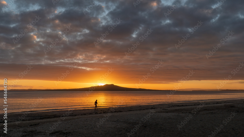 Silhouette woman walking on Milford beach. Sun rising over Rangitoto Island. Auckland.