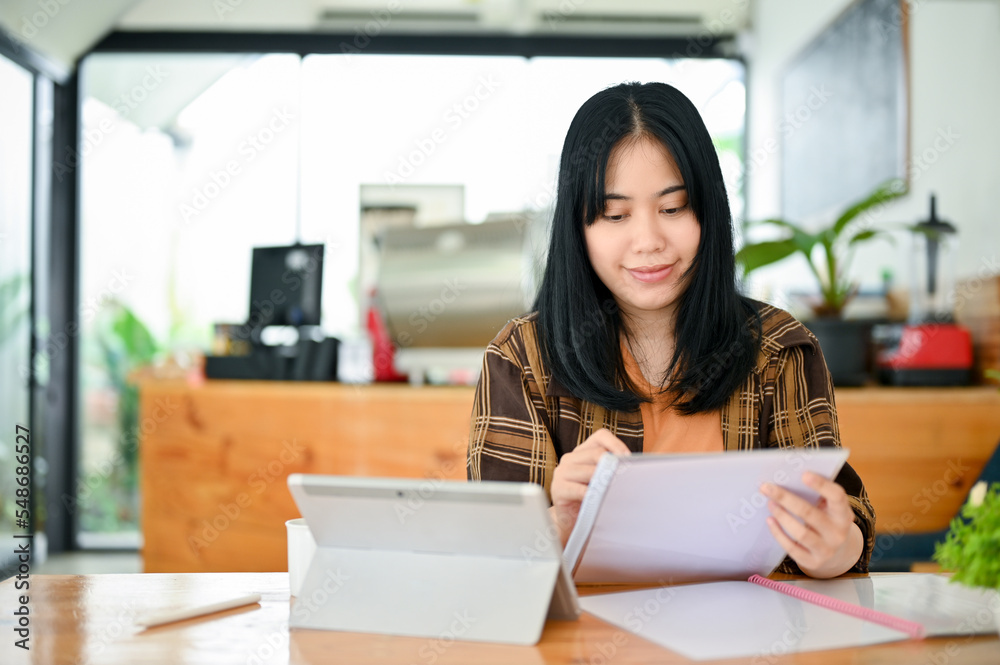 Fototapeta premium Charming Asian female remote working in the coffee shop, analyzing something on paperwork.