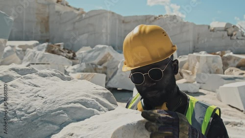 Bearded African American man in workwear and sunglasses checking quality of natural white marble stone extracted in quarry photo