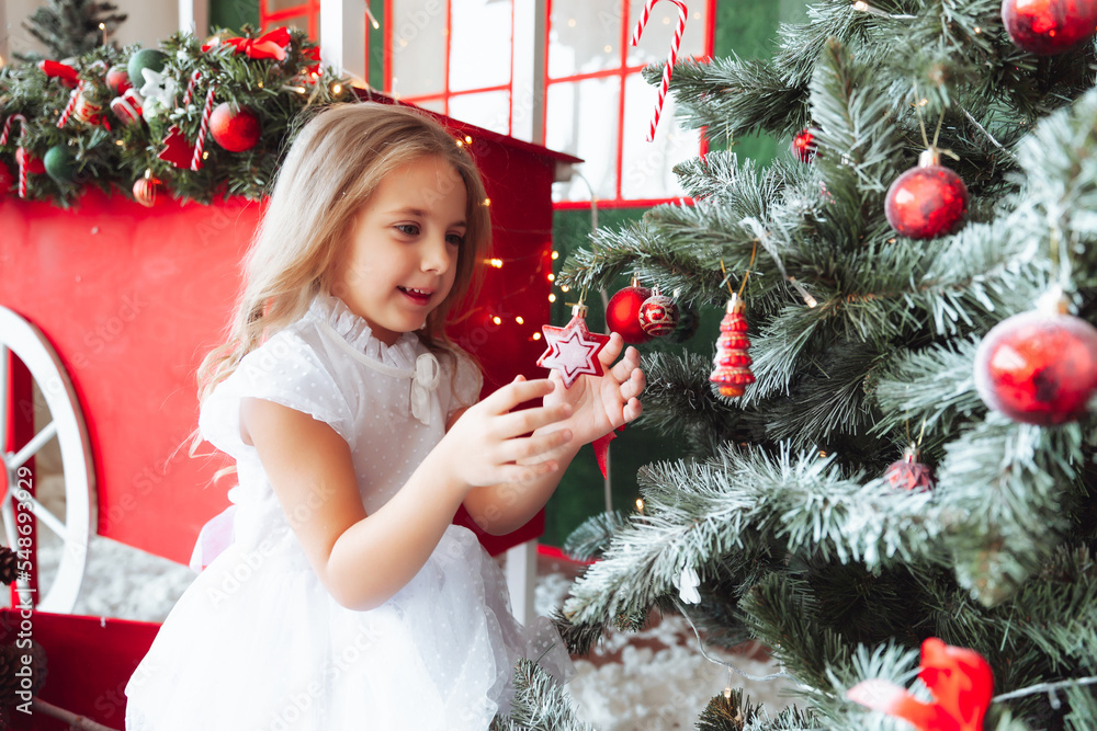 A little charming girl in a dress hangs a shiny ball on the Christmas tree. A child is decorating a Christmas tree in a beautiful living room. The concept of a happy family holiday New Year
