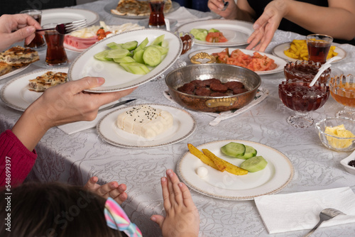 Family eating breakfast, over shoulder view of family eating breakfast. Little girl waiting for cucumber. Mother giving food. Friends having a good start to day. Cheese, sausage, fried potatoes, jar.