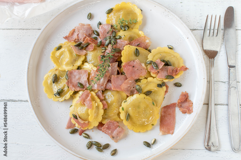 Ravioli with mushroom fillling, Cooked with italian ham, butter and roasted pumpkin seeds. Served on a white plate isolated on light wooden background. Ravioli with mushroom fillling, Cooked with italian ham, butter and roasted pumpkin seeds. Served on a white plate isolated on light wooden background.