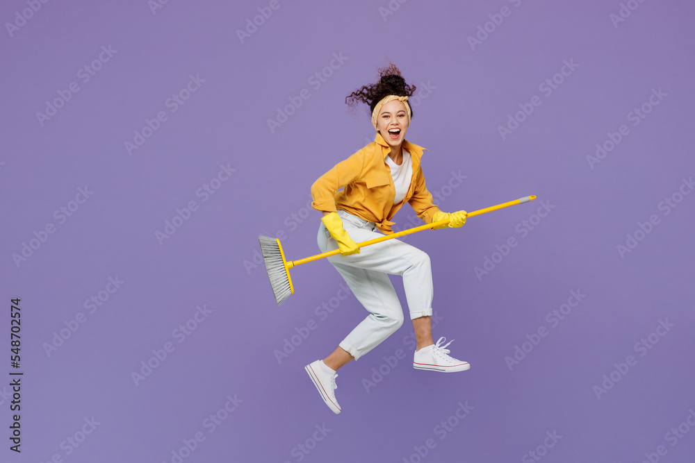 Full body side view smiling young housekeeper woman wear yellow shirt tidy up jump high hold broom sweep floor look camera isolated on plain pastel light purple background studio. Housework concept.