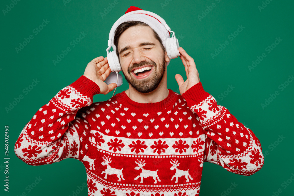 Merry relaxed satisfied young man 20s wear red warm knitted sweater Santa hat headphones posing listen music isolated on plain dark green background studio. New Year 2023 holiday celebration concept. Merry relaxed satisfied young man 20s wear red warm knitted sweater Santa hat headphones posing listen music isolated on plain dark green background studio. New Year 2023 holiday celebration concept.