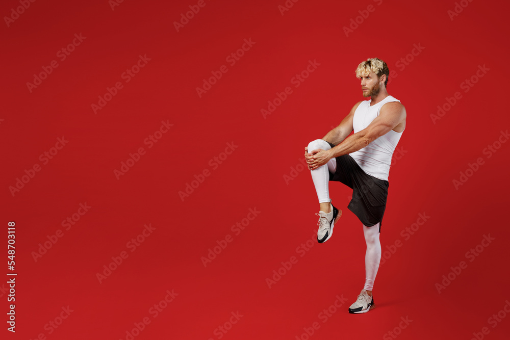 Full size side view young strong sporty sportsman man wear white clothes spend time in home gym train doing stretch exercise for legs isolated on plain red background. Workout sport fit body concept.