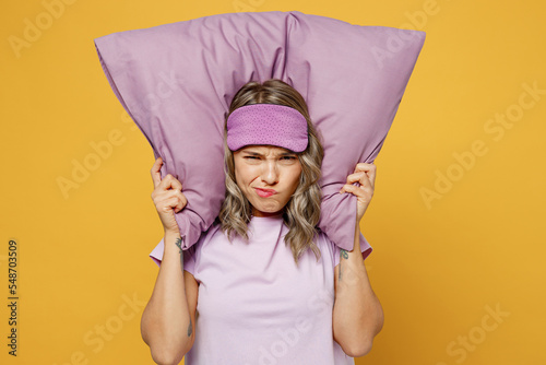 Sad tired young woman she wearing purple pyjamas jam sleep eye mask at home covering ears from neighbours noise snoring isolated on plain yellow background studio portrait. Bad mood night nap concept.